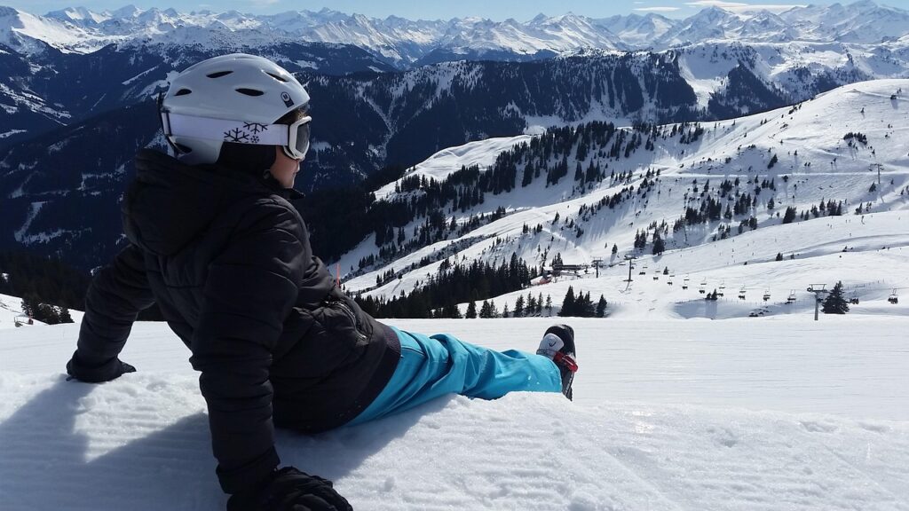 Esquiador sentado na neve, de capacete, a olhar para as montanhas dos Alpes